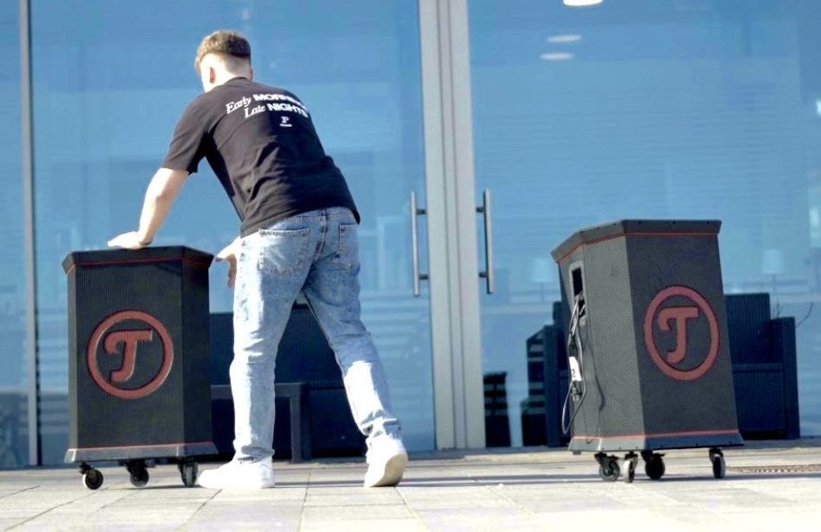 Soundanlage Person schiebt zwei schwarze Soundanlagen mit rotem Logo vor einem gläsernen Gebäude.